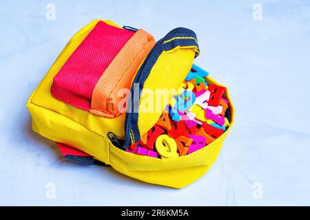 Essentiels pour la petite école : sac à dos miniature vibrant rempli de lettres en bois colorées isolées sur une table en béton. Banque D'Images