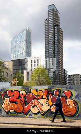 Londres, Royaume-Uni - 17 mai 2023: Un homme d'affaires en costume marche à côté d'un mur couvert de graffiti à Shoreditch, engrosé dans son téléphone, avec les grands bâtiments de Banque D'Images