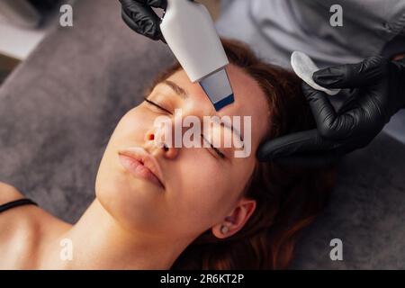 Maître de beauté fait peeling facial ultrasonique à son client. Jeune femme caucasienne faisant un nettoyage efficace de la peau et des pores dans un salo de beauté Banque D'Images