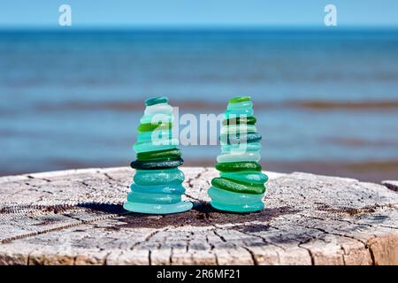 Deux pyramides équilibrées de fragments humides et polies de la mer d'une bouteille en verre bleu et vert sur un tronc en bois abîmé coupé contre la mer Banque D'Images