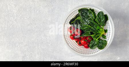Feuilles vertes lavées d'épinards et de tomates cerises sur une branche dans une passoire blanche en plastique sur une table en pierre légère. Vue de dessus avec espace de copie Banque D'Images