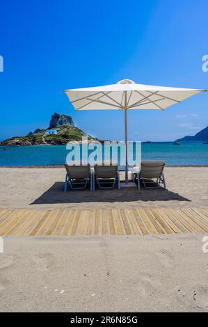 Chaise longue et parasol sur la belle plage d'Agios Stefanos en face de l'île Paradise Kastri - ruines historiques et paysage paradisiaque sur la côte de l'île Banque D'Images