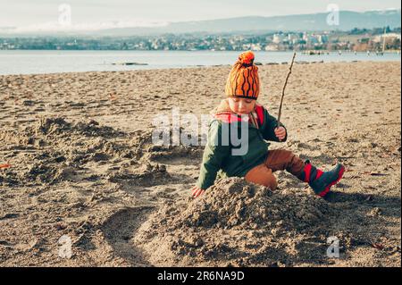 Adorable petit garçon jouant au bord du lac à l'extérieur, portant une veste chaude et des bottes d'hiver, bonne enfance Banque D'Images