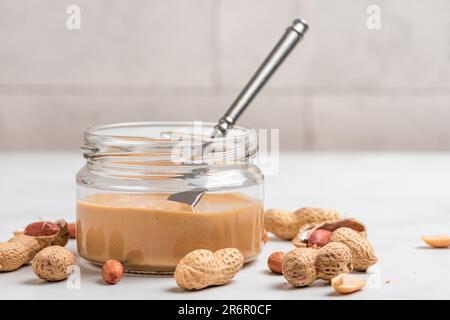 Beurre d'arachide dans un pot en verre, arachides, couteau de cuisine sur fond blanc. Nourriture végétalienne saine Banque D'Images