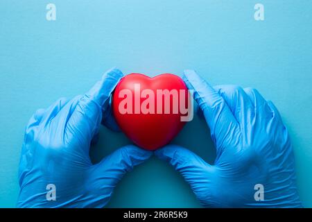 Main de professionnel médical portant des gants bleus protégeant un cœur rouge sur un fond bleu clair Banque D'Images