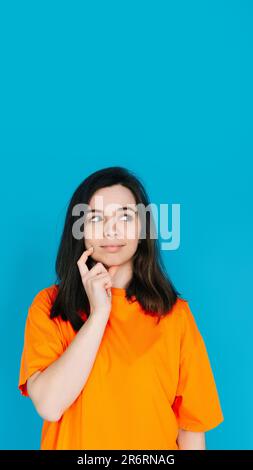 Jeune femme attrayante dans des vêtements orange à la mode, expression Pensive et sceptique, espace vide pour la copie, isolé sur fond bleu - Portrait photo Banque D'Images