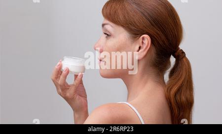 Dos de la pancaise pancaucasienne de taille moyenne visage de femme avec des taches de rousseur tenant ouvert bocal avec crème de peau à la main sur fond blanc Banque D'Images