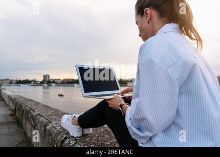 Femme urbaine assise sur le front de mer de la ville et utilisant son ordinateur portable, se rencontrant à la fin de la journée de travail hors du bureau. Banque D'Images