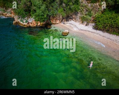 Femme se baignant et nageant sur la côte du Péloponnèse en eau verte et cristalline sur une plage romantique avec des rochers et une forêt en arrière-plan Banque D'Images