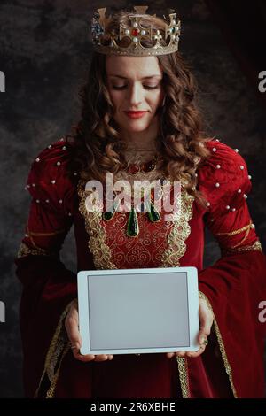 reine médiévale en robe rouge avec tablette numérique couronne écran vierge sur fond gris foncé. Banque D'Images