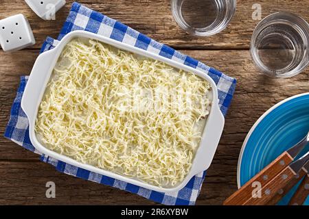 Lasagnes maison non cuites au four ou plat de cocotte avec fromage râpé et sauce au béchamel ou blanc sur le dessus, verres d'eau, assiettes avec fourchettes sur le côté Banque D'Images