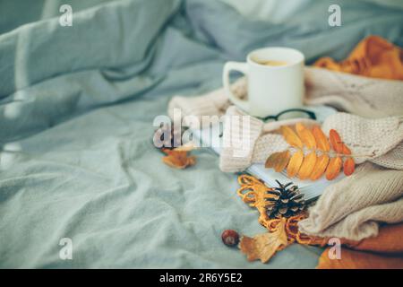 Ambiance chaleureuse. Chandail beige chaud, mug avec thé ou café, livre, lunettes et feuilles d'orange sur lit gris dans la chambre. Concept d'hygge. Banque D'Images