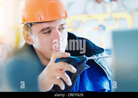 Un électricien ou un monteur d'instrumentation muni d'un tournevis à la main règle le fonctionnement d'un manomètre dans une installation industrielle de gaz intérieure. Travailleur caucasien dans un casque de sécurité orange et un pelage de pois d'hiver au travail. Portrait en gros plan. Banque D'Images