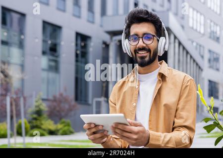 Portrait d'un jeune Indien avec un casque et une tablette regardant des vidéos en ligne assis sur un banc près d'un homme d'affaires de bâtiment de bureau souriant et regardant l'appareil photo, portrait d'un lecteur de flux en ligne satisfait Banque D'Images