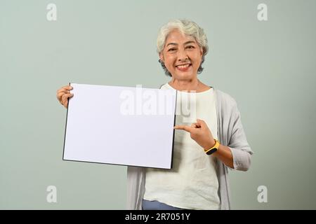 Portrait d'une femme souriante aux cheveux gris 60s tenant un tableau publicitaire vierge debout sur fond bleu clair Banque D'Images