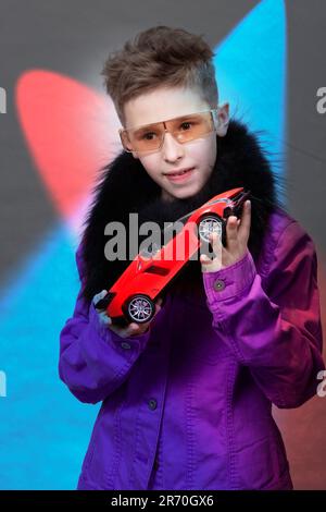 Un garçon à la mode tient une voiture-jouet dans ses mains, sur un fond de studio multicolore. Un bel enfant dans des verres futuristes rêvant d'une exte Banque D'Images