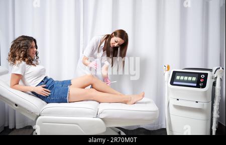 Un cosmétologue souriant en gants stériles applique du gel sur la jambe avant l'épilation. Jeune femme allongé sur un lit de jour près de l'appareil d'épilation à diode laser tout en ayant une procédure d'épilation en clinique. Banque D'Images