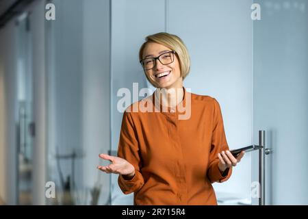 Portrait d'une femme d'affaires joyeuse à l'intérieur du bureau sur le lieu de travail, l'économiste comptable regarde avec joie l'appareil photo et tient un smartphone entre ses mains. Banque D'Images