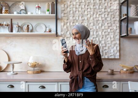 Une jeune femme musulmane agréablement choquée dans le hijab debout dans la cuisine et regardant le téléphone. Se réjouit, crient avec plaisir, lit des nouvelles, des messages. Banque D'Images