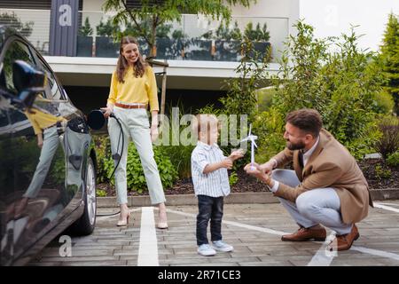 Une famille heureuse se tenant à côté de sa voiture et se chargeant de la voiture électrique dans la rue. Banque D'Images