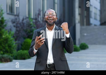 Portrait réussi et satisfait patron d'homme d'affaires afro-américain tient le téléphone dans les mains souriant et regardant l'appareil-photo, l'homme a reçu la notification de la victoire réussie, tient la main pour célébrer le triomphe Banque D'Images