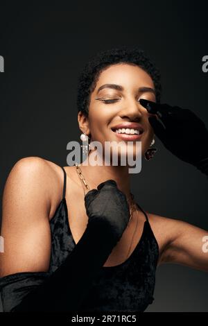 Portrait de femme afro-américaine à la mode et souriante en robe et gants touchant le visage tout en fermant les yeux et en se tenant isolé sur noir, haut f Banque D'Images