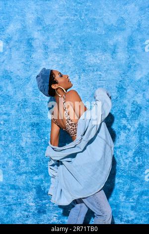 Vue latérale de la femme afro-américaine en béret, haut à imprimé animal et chemise en denim posé et debout sur fond bleu texturé, styli Banque D'Images