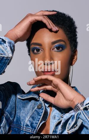 Portrait d'une femme afro-américaine moderne avec cheveux courts et maquillage audacieux touchant le visage et regardant l'appareil photo tout en posant dans une tenue en denim isolée sur Banque D'Images