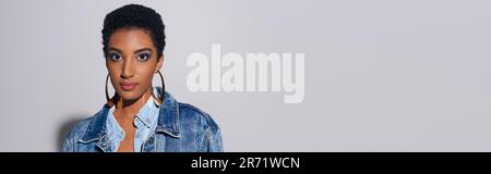 Portrait du jeune modèle afro-américain à la mode avec un maquillage audacieux dans une veste en denim et des boucles d'oreilles dorées regardant l'appareil photo sur fond gris avec co Banque D'Images