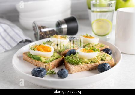 Pain grillé avec tartiner à l'avocat, œufs durs et cresson sur une assiette. Servi avec un verre d'eau et du café sur une table de cuisine. Banque D'Images