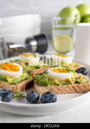 Pain grillé avec tartiner à l'avocat, œufs durs et cresson sur une assiette. Servi avec un verre d'eau et du café sur une table de cuisine. Banque D'Images