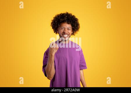 Gai noir adulte curly homme dans le t-shirt violet fait le geste de la victoire, célèbre le succès Banque D'Images