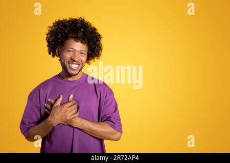 Gai noir adulte homme curly dans le t-shirt violet fait le geste de gratitude, mettre les mains sur la poitrine Banque D'Images