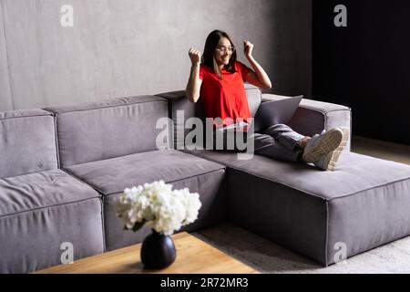 Une jeune femme surjoyeuse s'assoit sur un canapé à la maison triomphe recevoir un e-mail agréable ou lire de bonnes nouvelles sur un ordinateur portable Banque D'Images