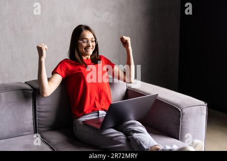 Une jeune femme surjoyeuse s'assoit sur un canapé à la maison triomphe recevoir un e-mail agréable ou lire de bonnes nouvelles sur un ordinateur portable Banque D'Images