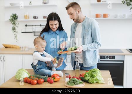 Un père choqué tient une planche à découper avec des tranches de concombre, tandis qu'une petite fille douce mélange la salade avec les mains dans un bol en verre. Curieux enfant explorant la texture et les formes de punch coloré des ingrédients de la salade. Banque D'Images