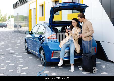 Les jeunes amis chargent la batterie de l'automobile à partir de la gare publique et se tiennent près du parking pour la voiture électrique. Homme et femme parlant et buvant du café Banque D'Images