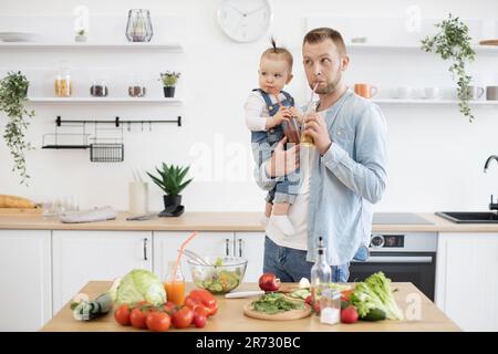 Le père et la fille boivent des boissons biologiques saines à travers des pailles en plastique et regardent de côté. Papa et fille caucasien aidant à préparer un dîner savoureux dans la cuisine moderne blanche. Banque D'Images