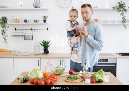 Le père et la fille boivent des boissons biologiques saines à travers des pailles en plastique et regardent de côté. Papa et fille caucasien aidant à préparer un dîner savoureux dans la cuisine moderne blanche. Banque D'Images