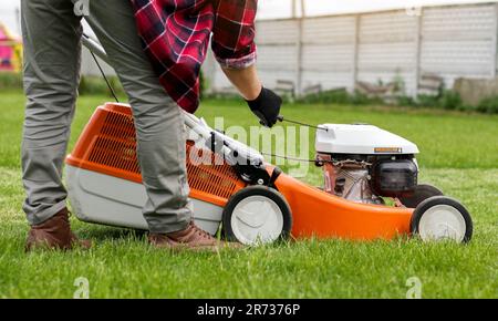 Un jardinier méconnaissable en gants de protection démarre le robot de tonte avant de couper de l'herbe verte dans son jardin. Homme avec tondeuse à gazon motorisé env Banque D'Images