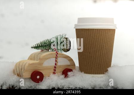 Voiture en bois portant l'arbre de Noël avec une tasse en papier maquette de café ou de chocolat chaud sur la neige. Espace de copie pour le texte voiture miniature dans un paysage enneigé. Joyeux Noël et bonne année concept. Carte de vœux pour l'hiver à venir Banque D'Images