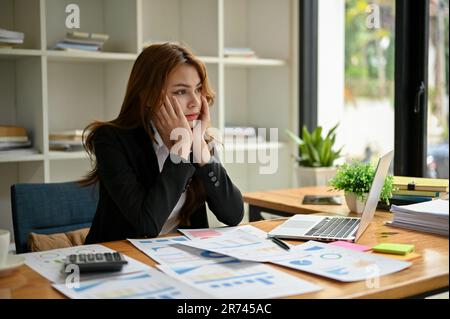 Une jeune femme d'affaires ou une travailleuse financière asiatique frustrée et ennuyeuse s'assoit à son bureau, les mains sur le menton, en regardant par la fenêtre avec un Banque D'Images