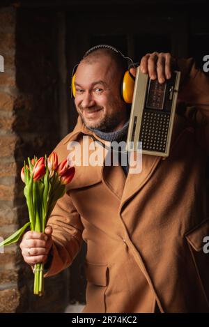 Regard drôle d'un homme avec un bouquet, une radio rétro et un casque dans un manteau. Une image absurde pour une date. Banque D'Images