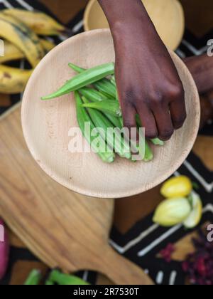 une femme africaine hache des légumes aux fruits d'okra pour cuisiner une recette traditionnelle avec d'autres légumes et fruits locaux. Banque D'Images