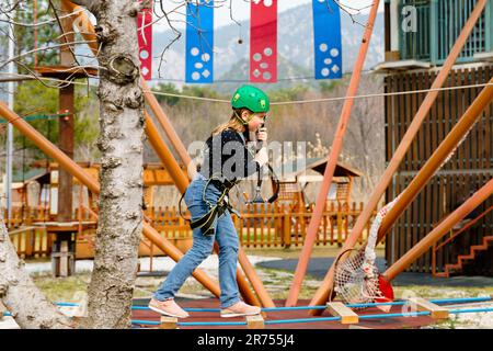 Adolescente dans l'équipement de harnais d'escalade, casque de sécurité vert de sport. Parc d'attractions de corde. Fixation fixant le mousqueton à la corde de sécurité. Hangin Banque D'Images