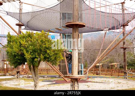 Adolescente dans l'équipement de harnais d'escalade, casque de sécurité vert de sport. Parc d'attractions de corde. Fixation fixant le mousqueton à la corde de sécurité. Hangin Banque D'Images