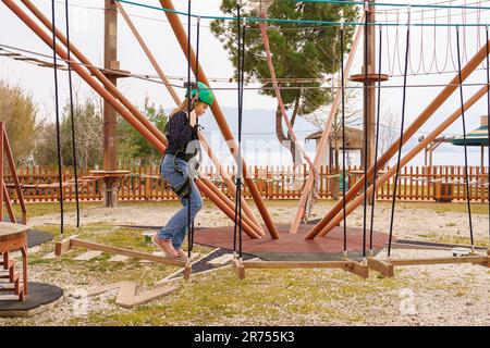 Adolescente dans l'équipement de harnais d'escalade, casque de sécurité vert de sport. Parc d'attractions de corde. Fixation fixant le mousqueton à la corde de sécurité. Hangin Banque D'Images