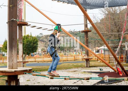 Adolescente dans l'équipement de harnais d'escalade, casque de sécurité vert de sport. Parc d'attractions de corde. Fixation fixant le mousqueton à la corde de sécurité. Hangin Banque D'Images