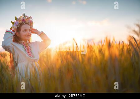 Belle jeune fille avec un chapelet de fleurs, robe folklorique ethnique avec broderie bulgare traditionnelle pendant le coucher du soleil sur un champ agricole de blé Banque D'Images