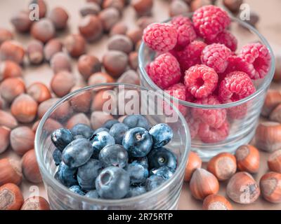 Gros plan de framboises mûres fraîches et de bleuets dans des verres peu transparents et de noisettes fermées sur fond flou. Banque D'Images